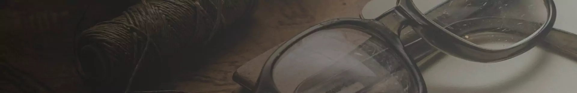 Banner de um óculos de grau com a armação quadrada na cor preta e marrom sobre uma mesa com um rolo de linha ao lado