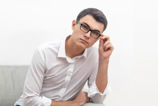 Homem com cabelo preto sentado em um sofá usando um óculos de grau quadrado com armação preta